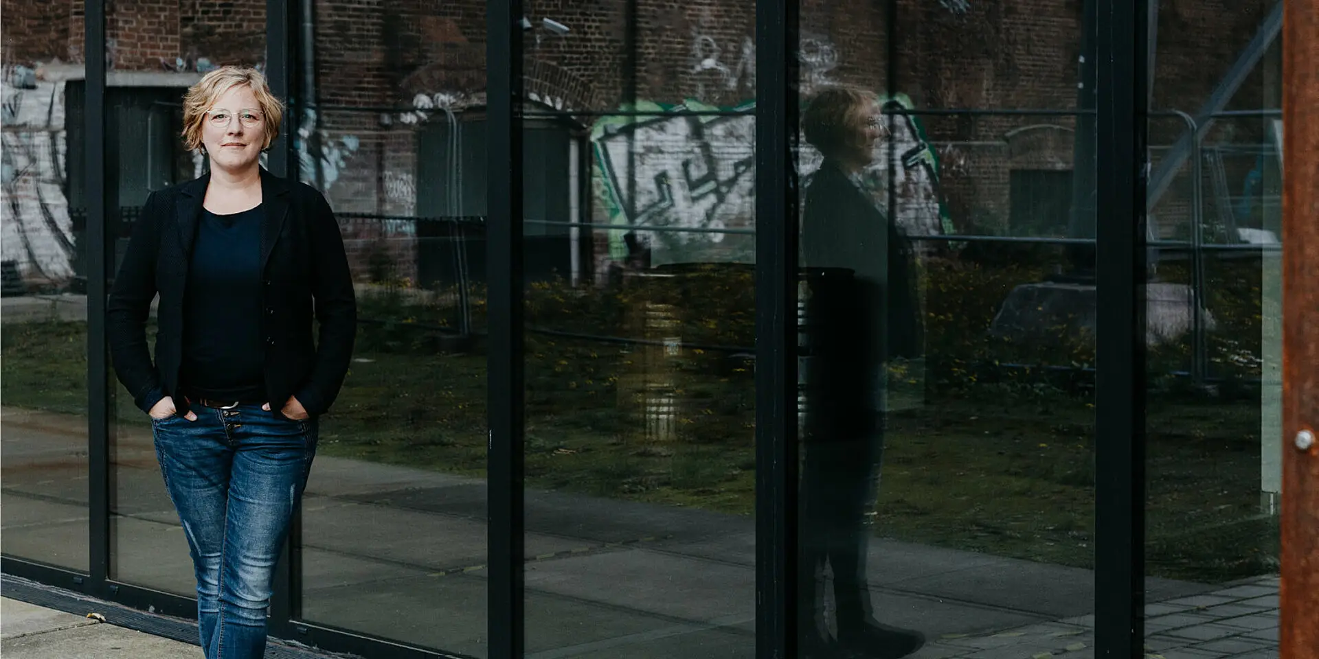 Katharina Schulte für einer sich spiegelnden, modernen Glasfassade. Im Hintergrund die Mauern einer alten Industrieanlage